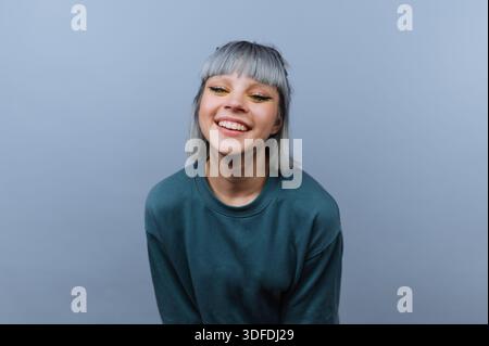 A young woman sits with a big smile on her face. She has short gray hair and wears a green shirt. The background is gray with good lighting making her Stock Photo