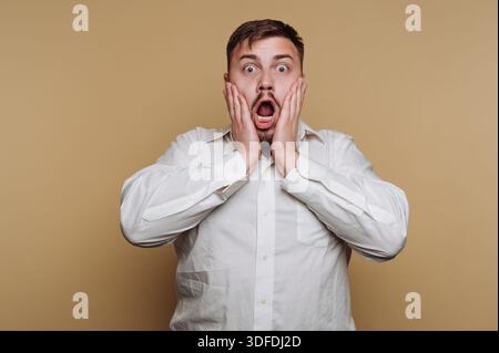 A man stands in a studio with a neutral background, showing surprise with his mouth open and hands on his cheeks. He appears shocked in the bright lig Stock Photo