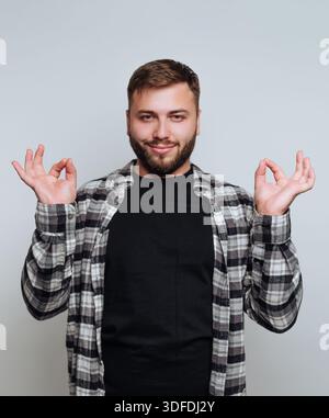 A man stands in front of a gray background with a relaxed expression. He raises his hands to form a gesture often associated with meditation. His outf Stock Photo