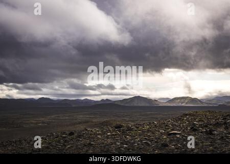 View of a stark, volcanic landscape unfolds under a dramatic sky, where dark clouds loom over distant mountains, creating a scene of raw beauty, Landm Stock Photo