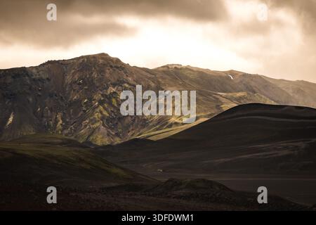 View of jagged, dark mountains rise against a stormy sky, their slopes marked with streaks of green, contrasting with the shadowed foreground, Landman Stock Photo