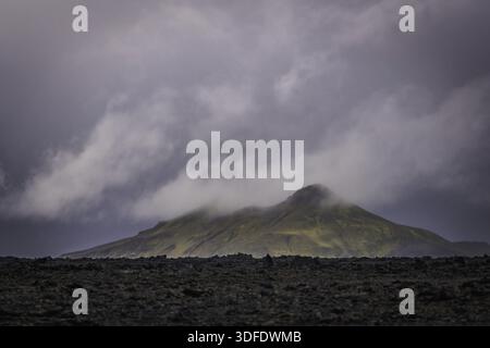 View of a lone peak veiled in mist rises dramatically from a field of black volcanic rock under a sky heavy with brooding clouds, Landmannalaugar, The Stock Photo