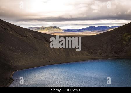 View of a serene blue lake nestled between dark, volcanic slopes under a cloudy sky, creating a stark contrast in the Icelandic highlands, Landmannala Stock Photo