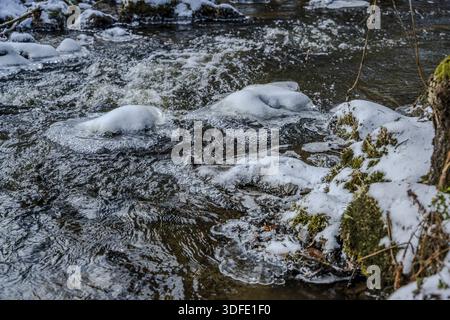Water flows through a forest with rocks and snow on the shore. The drops of water are splashing and there are some snow-covered areas. Hoellbachtal, U Stock Photo