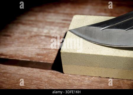 Knife blade resting on a whetstone block, getting sharpened on a wooden background. Maintenance tool for sharp edge Stock Photo