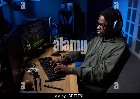 Young Black Man Wearing Headphones Coding on Computer in Dark Office Stock Photo