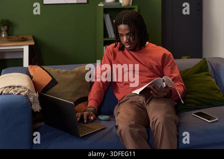 Young Black Man Studying with Notebook and Laptop While Sitting on Sofa Stock Photo