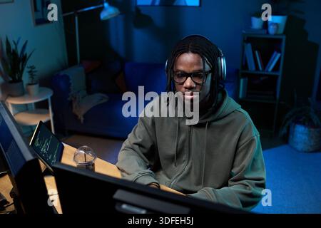 Young Black Man Wearing Headphones Coding on Computer in Home Office at Night Stock Photo