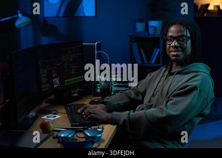 Portrait of Young Black Man Coding on Computer at Desk in Dark Office Stock Photo