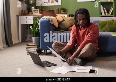 Young Black Man Sitting on Floor Reviewing Documents and Using Laptop Stock Photo