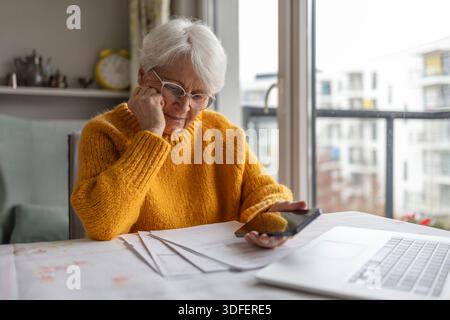Senior woman holding mobile phone while reading paper documents at home Stock Photo