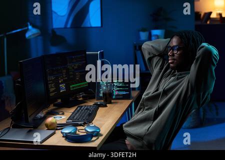 Young Black Man Relaxing While Coding on Computer Monitors at Night Stock Photo