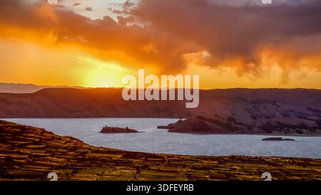 View of a sunset on Lake Titicaca with a golden sky reflecting on the water, small islands dotting the horizon, and terraced fields in the foreground, Stock Photo