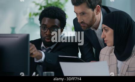 Three multiracial business colleagues in office with computer and paper document discuss startup project looking at screen multiethnic diversity Stock Photo