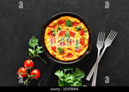 Frittata with tomatoes and spinach in frying pan over black stone background. Top view, flat lay Stock Photo