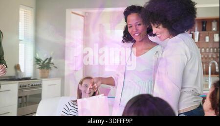 Smiling two women holding pale pink gift bag in modern living area, wearing light dresses Stock Photo