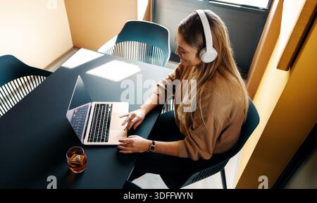 A young adult woman works on her laptop while enjoying music with headphones, creating a focused and modern setting. A relaxed and professional enviro Stock Photo