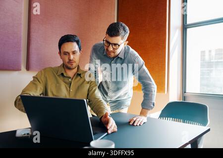 Two men in a modern office environment, engaged in discussions while working on a laptop, demonstrating teamwork and a collaborative professional atmo Stock Photo
