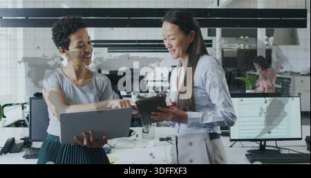 Collaborating two women wearing office attire holding laptop and tablet at office with map decal Stock Photo