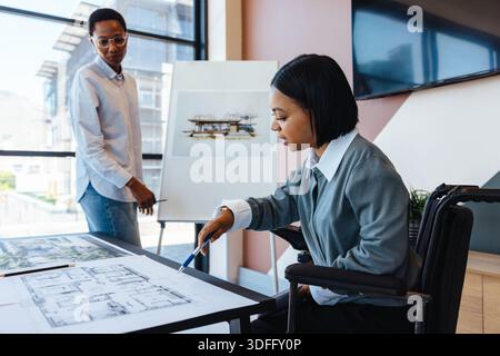 Two women working on design plans in an office, one seated in a wheelchair. The scene portrays professional collaboration, inclusive workspace, archit Stock Photo