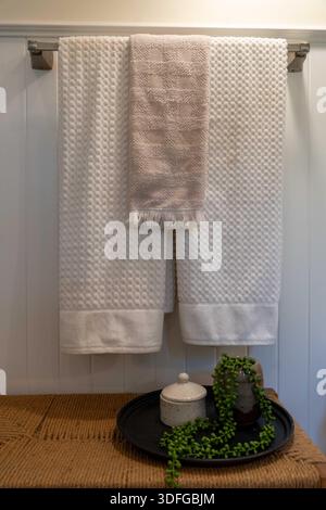 White towels hang neatly on a rustic rack. Small green plant rests beside a lidded jar. The scene evokes calm and order. Perfect for home decor. Stock Photo