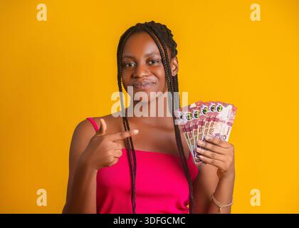 A young woman with braids smiles against a bright yellow background. She wears a pink top and points toward a fan of 200 Ghanaian Cedi banknotes held Stock Photo