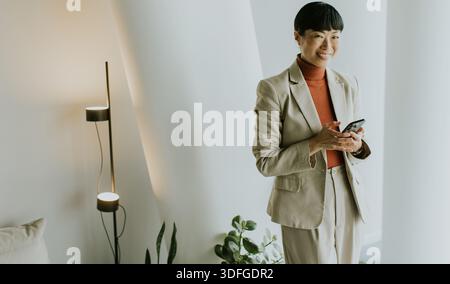 A businesswoman stands confidently in a light-colored suit, smiling while browsing her phone. She is in a modern workspace with indoor plants and soft Stock Photo