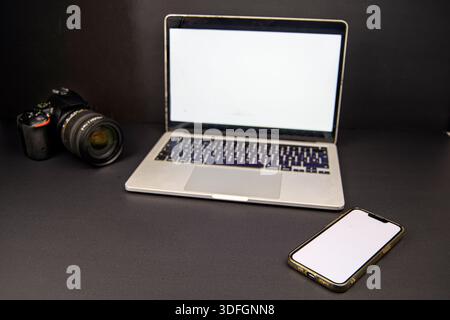 Dark minimal workspace with laptop and smartphone blank screens and DSLR camera on black background, copy space for modern tech message. Stock Photo