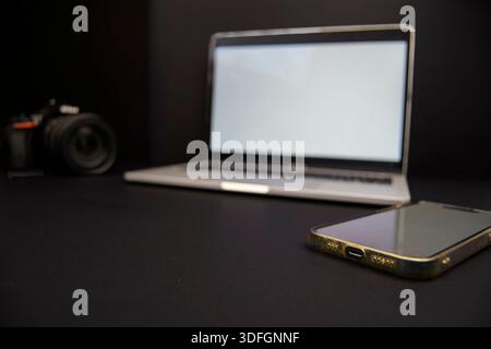 Smartphone close-up in foreground with laptop and DSLR camera blurred in dark background, concept of mobile workflow, communication and creative tech. Stock Photo
