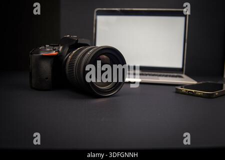 Dark creative workspace with DSLR camera, laptop and smartphone. Blank screens and copy space for content creation and remote work. Stock Photo