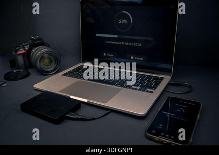 Laptop and smartphone showing file transfer progress with a camera and external hard drive. Backup, storage and data management workflow. Stock Photo