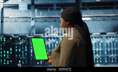 Data center specialist monitors data center metrics on green screen tablet to enhance operational effectiveness. Server room worker uses analytics on chroma key device to predict performance, camera B Stock Photo