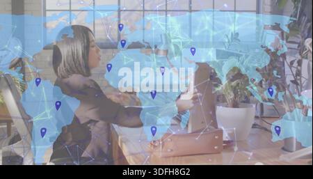 Working Japanese woman in blazer leaning at office desk, using laptop and binder, world map overlay Stock Photo