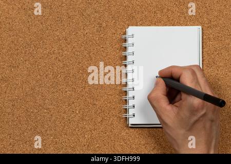 Close up to a male hand ready to writing into an a small white notebook at the left side of the screen over a cork background Stock Photo