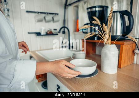 woman hands places a white bowl on a wooden table in a cozy kitchen. A vase with dried wheat and a coffee maker are visible. Stock Photo