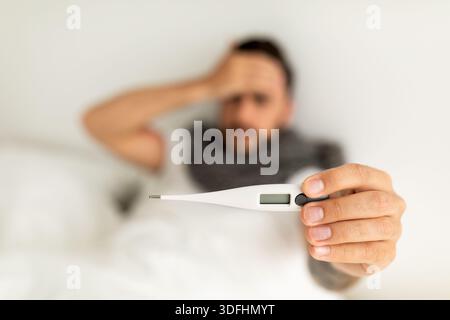 Sick young man showing thermometer to camera, lying on bed and touching forehead. Selective focus, top view Stock Photo
