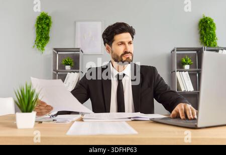 Confident businessman in suit reviewing documents at desk and working on laptop Stock Photo
