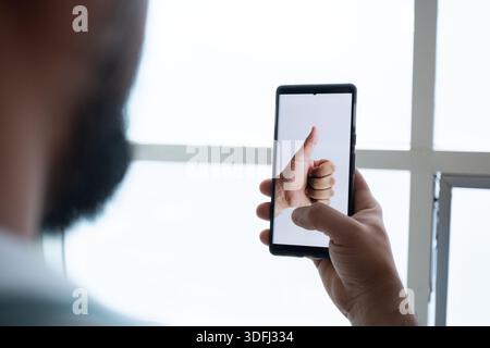 A person holding a smartphone displaying a thumbs up gesture, with a bright window in the background Stock Photo