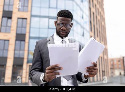 Young black man in suit checking reports or getting ready for job interview, holding documents or CV, standing outdoors Stock Photo