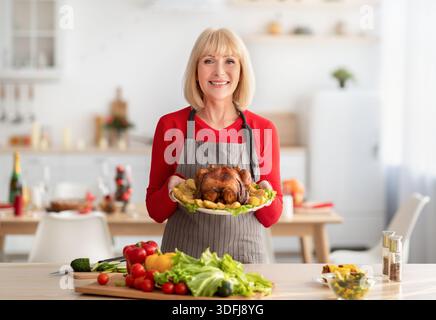 Cheerful elderly woman in apron holding dish with roasted turkey for Christmas or Thanksgiving holiday in kitchen Stock Photo