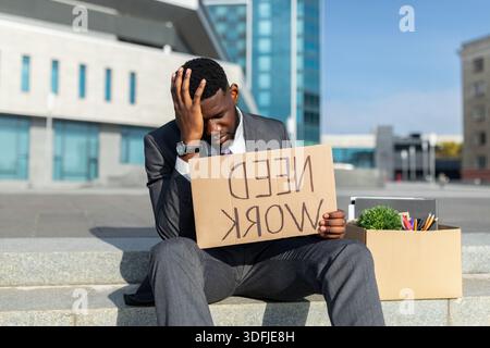Fired african american businessman with paper poster saying need work and box of personal things, sitting outdoors Stock Photo