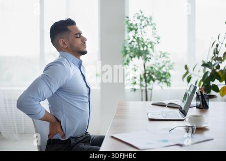 Young Arab businessman having acute back pain, massaging aching muscles, sitting at desk in modern office, copy space Stock Photo