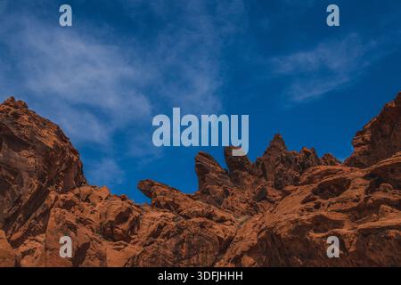 View of rugged, fiery-red rock formations reaching towards a vibrant blue sky dotted with wispy clouds, creating a striking contrast in Valley of Fire Stock Photo