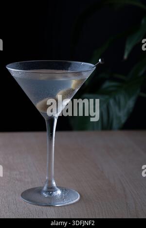 classic martini cocktail glass with two olives on a pick, photographed against a background of green leaves in a modern home bar setting Stock Photo
