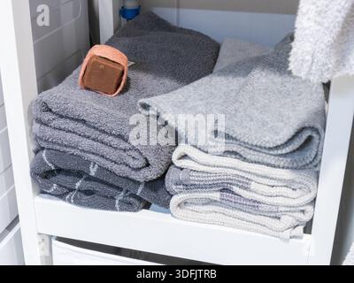 Folded grey and white towels are neatly stacked on a white shelf, with a brown, string-tied soap bar resting on the top left stack, suggesting organiz Stock Photo