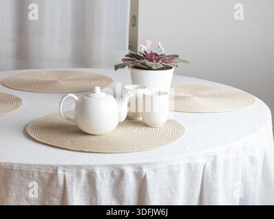 A ceramic teapot sits on a round placemat on a white-covered table, flanked by two white cups and a potted African violet plant. The scene suggests a Stock Photo