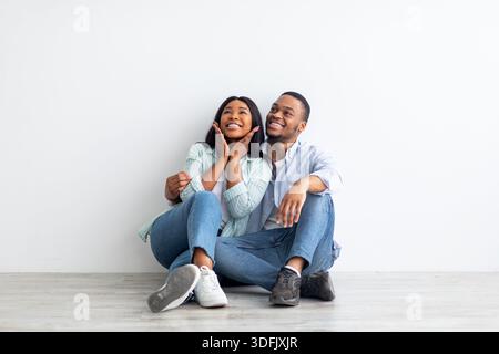 Dreaming about future. Excited black couple sitting on floor leaning on wall in new apartment, looking at free space Stock Photo