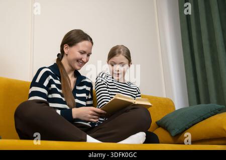 Happy mother and daughter spending quality time, sitting on a yellow sofa and reading a book, fostering learning and family connection in their home e Stock Photo