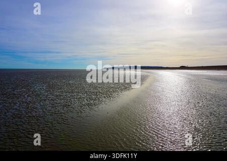 Low tide on a wide sandy beach in England, with shallow water reflecting the sky under soft sunlight, creating a calm coastal landscape. Stock Photo