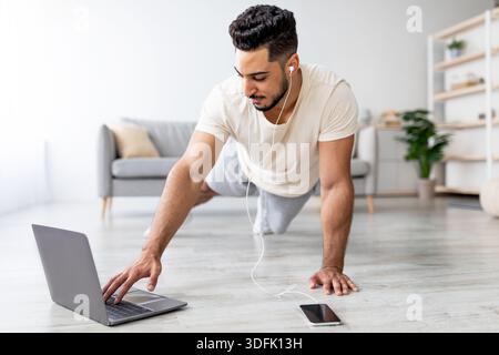 Young Arab guy doing plank, wearing headphones, listening to music, following online video tutorial on laptop at home Stock Photo